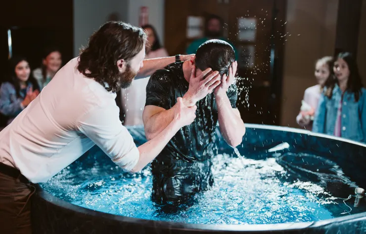 Man baptizing another man by immersing him in water inside a baptismal pool with people watching in the background.