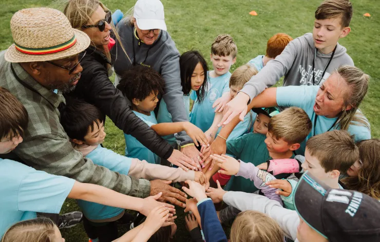 A diverse group of children and adults standing in a circle outdoors with their hands extended inward in a team huddle.