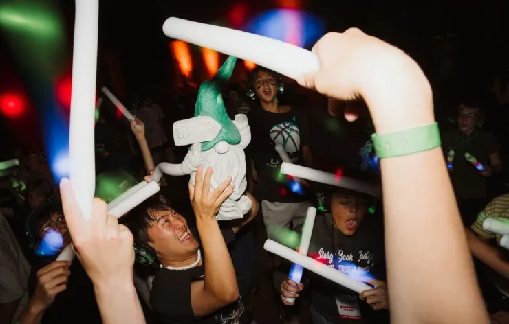 People dancing energetically in a dark room holding glowing light sticks, one person holding a garden gnome statue.