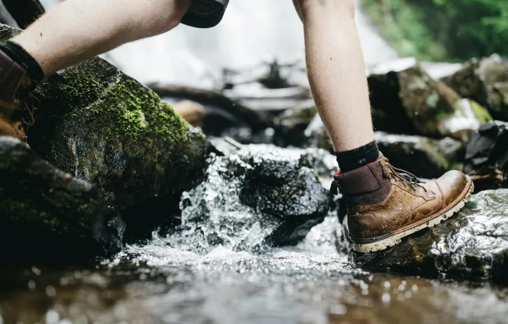 Person wearing brown hiking boots stepping on wet rocks while crossing a stream.