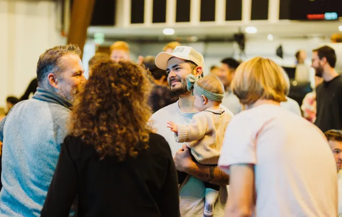 A man wearing a cap holding a baby with a bow headband, talking to three adults in a busy indoor setting.