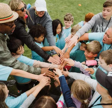 A diverse group of children and adults outdoors joining their hands together in a circle.