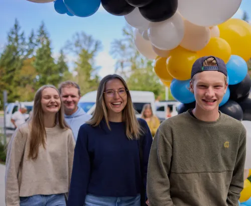 Four young adults smiling and walking outdoors under an arch of white, black, yellow, and blue balloons with trees and vehicles in the background.