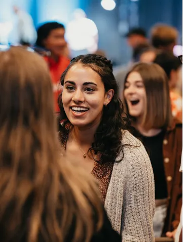 Smiling young woman with curly hair talking to another person at a social event with blurred people in the background.