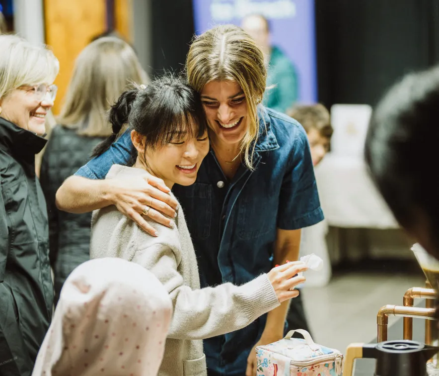 Two women smiling and hugging at a social gathering, with other people in the background.