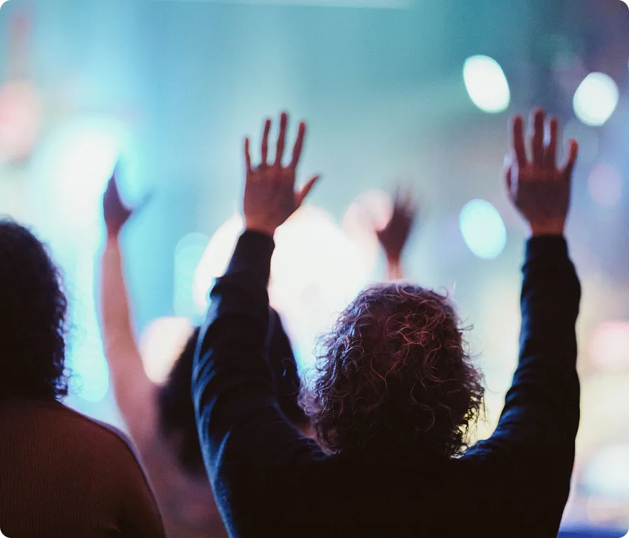 People raising hands in a dimly lit setting with colorful blurred lights in the background.