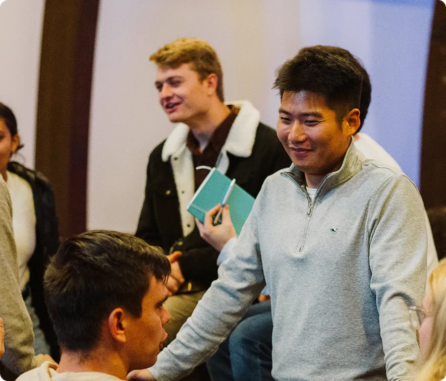 Group of young people engaged in conversation, one holding a notebook and pen.