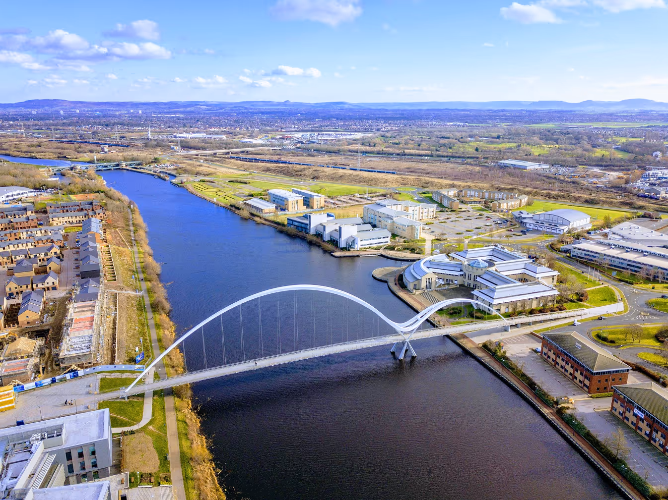 Aerial view of a modern arched pedestrian bridge spanning across a river with buildings and greenery on both sides under a partly cloudy sky.