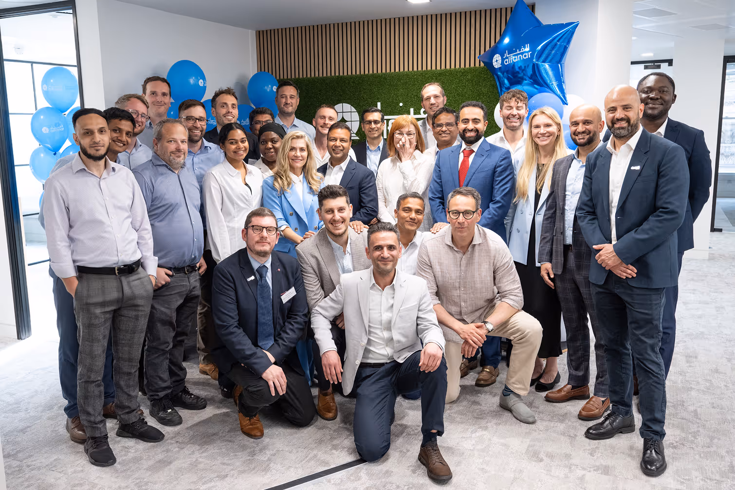 A diverse group of professionally dressed people posing together indoors with blue alfanar branded balloons in the background.