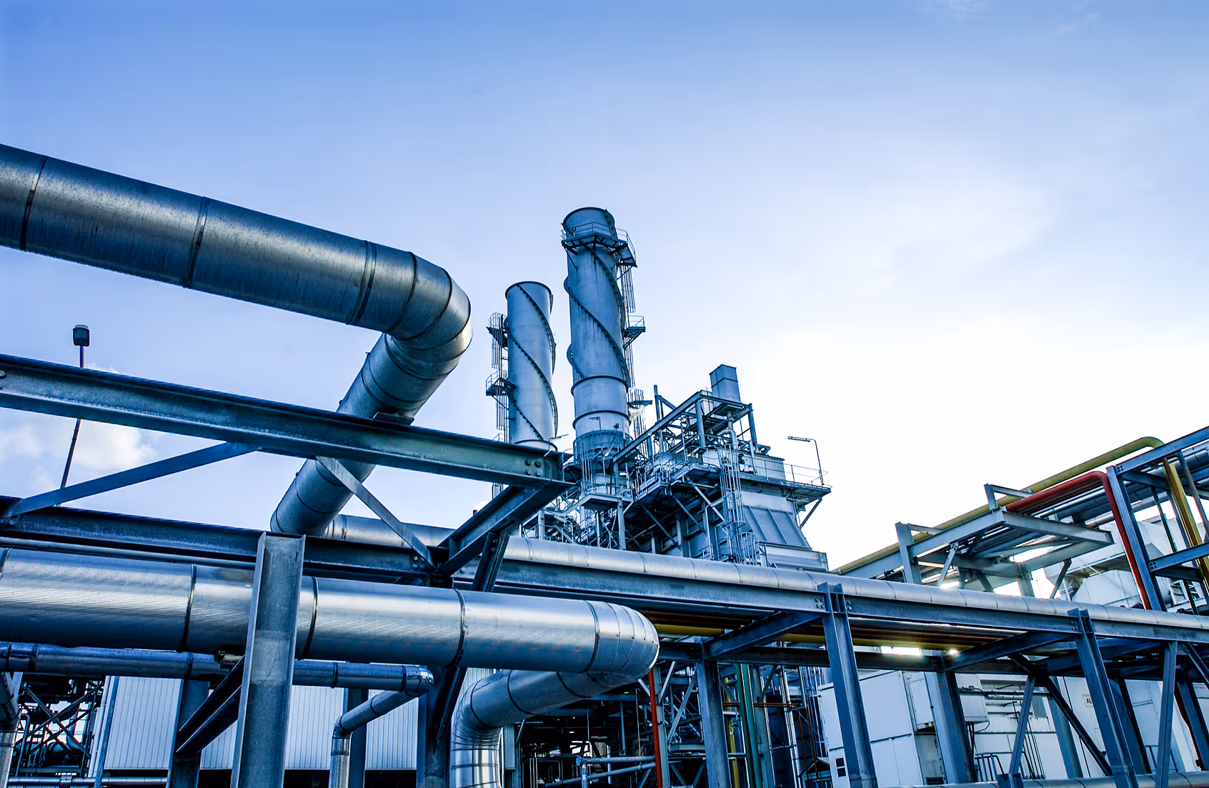 Industrial factory with large metal pipes and tall cylindrical chimneys against a blue sky.