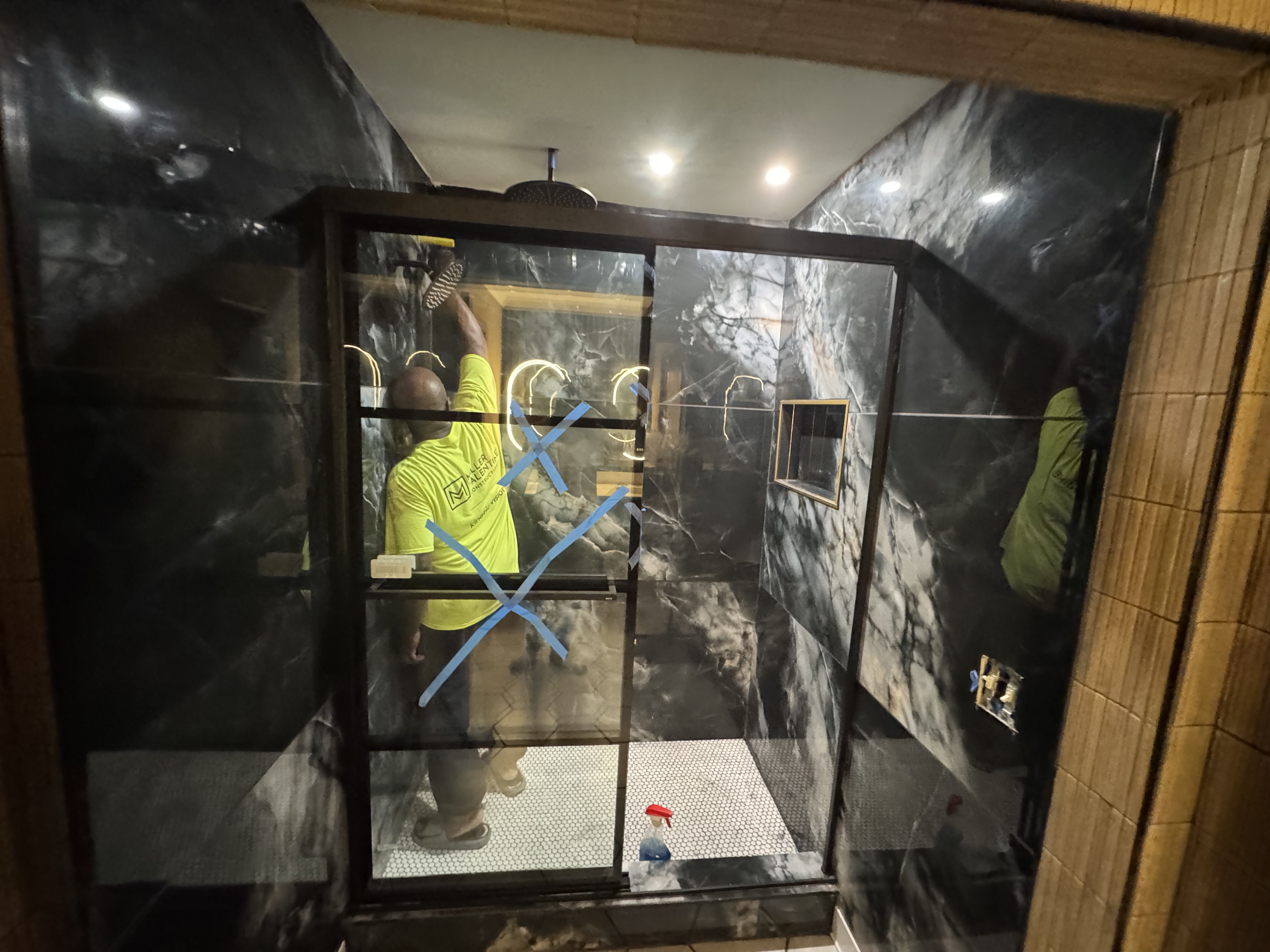 Man in a yellow shirt cleaning a modern black marble shower with glass door marked with blue Xs.