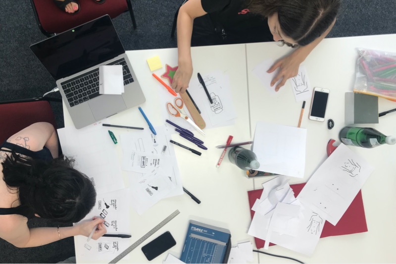 Aus der Vogelperspektive werden 2 Personen gezeigt, die an einem weißen Tisch mit Laptop, Stift und Papier sitzen.