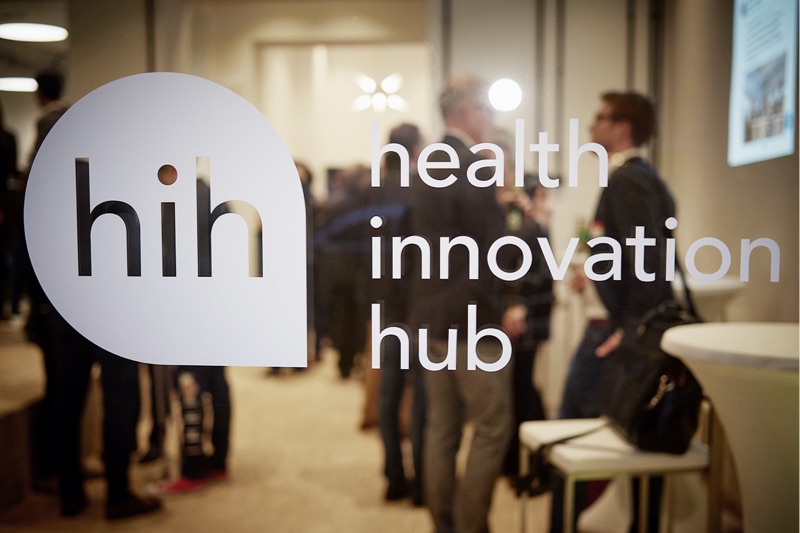 Glasscheibe mit dem Health innovation Hub Logo und Schriftzug. Im Hintergrund sind mehrere Personen.