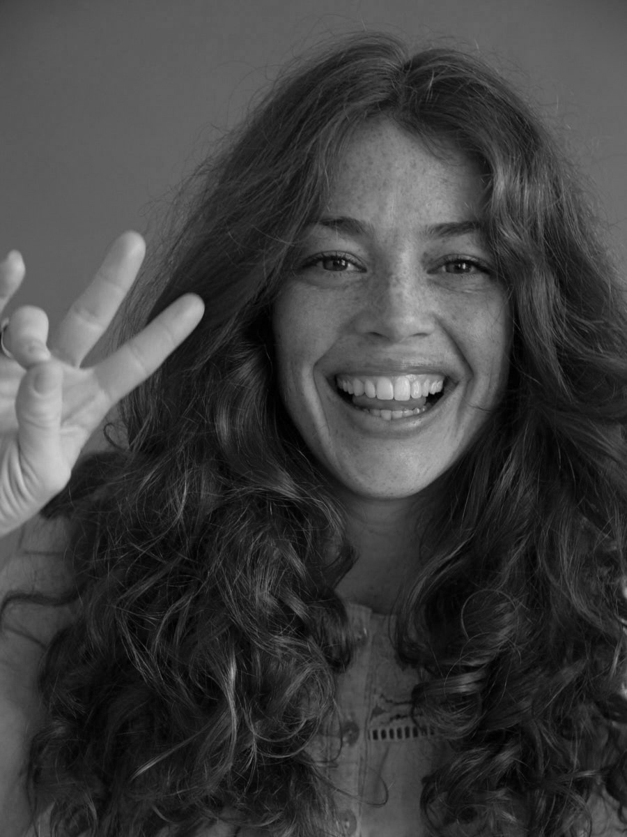 Black-and-white portrait of a smiling woman with curls and freckles, seen from the front, making a peace sign with her hand.