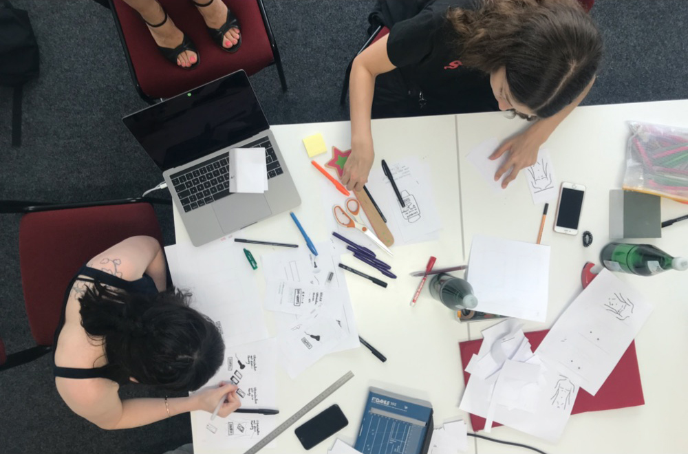 Aus der Vogelperspektive werden 2 Personen gezeigt, die an einem weißen Tisch mit Laptop, Stift und Papier sitzen.