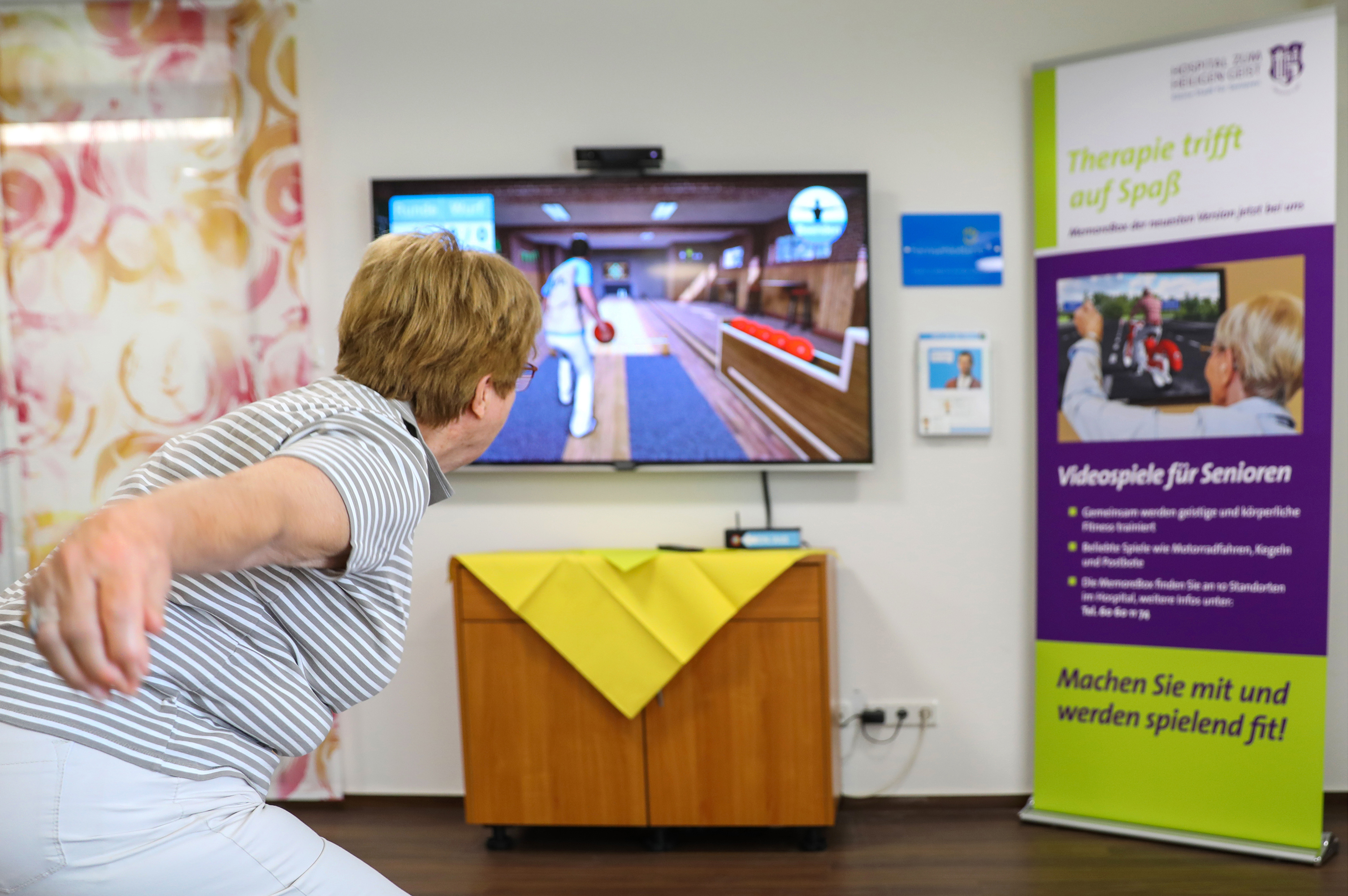 Eine Seniorin führt eine Wurfbewegung vor einem Bildschirm aus, auf dem eine Bowling-Simulation zu sehen ist.