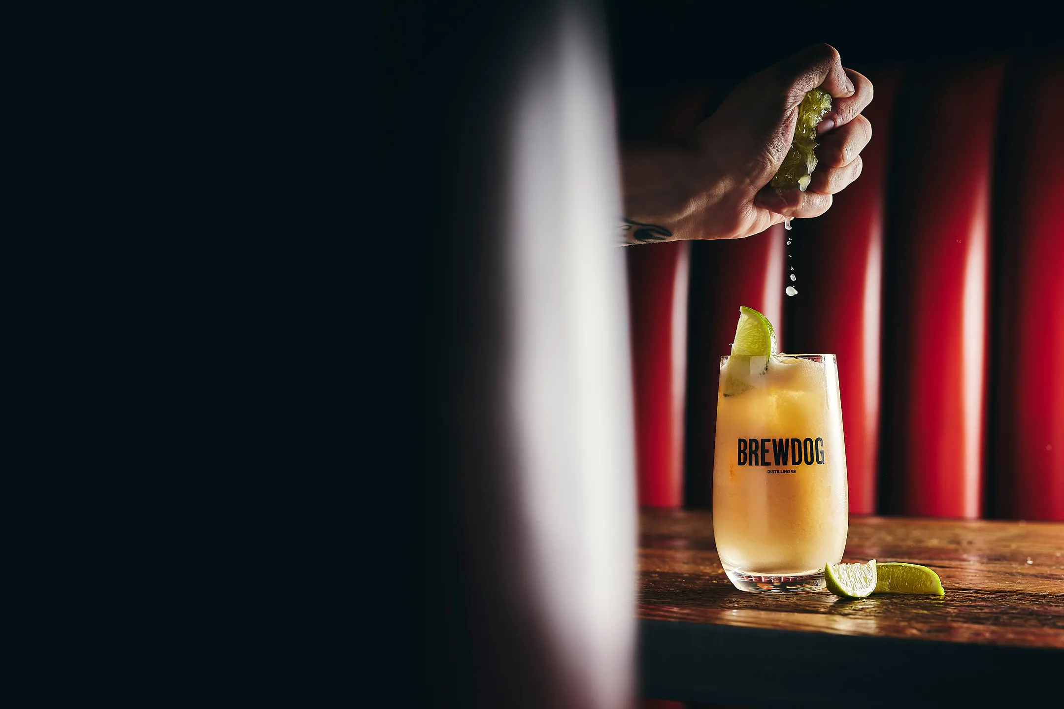 Hand squeezing a lime wedge over a BrewDog branded cocktail glass on a wooden table with lime slices and a red cushioned background.