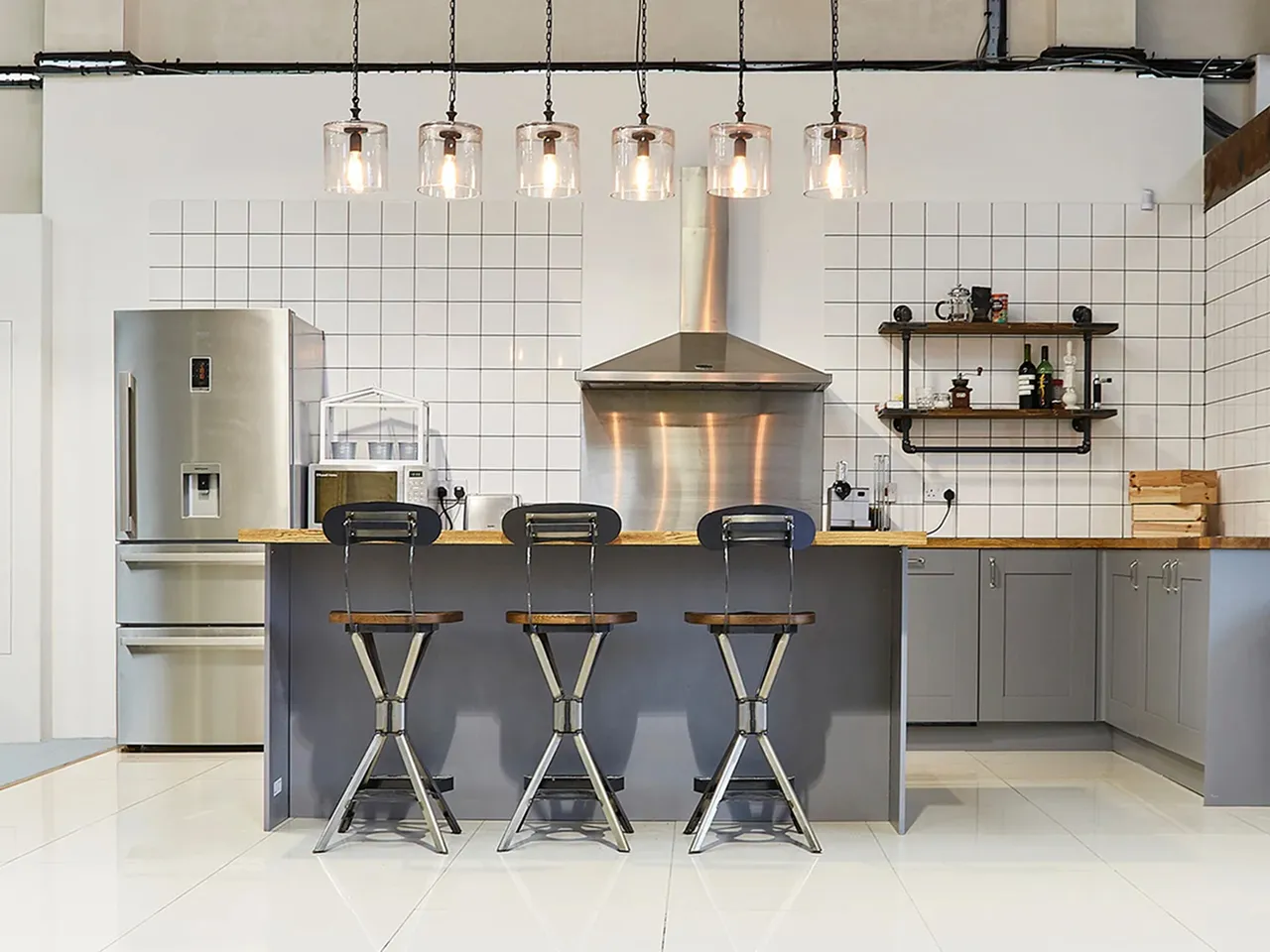 Modern kitchen with grey cabinets, stainless steel refrigerator, wood-topped island with three metal stools, and hanging glass light fixtures.