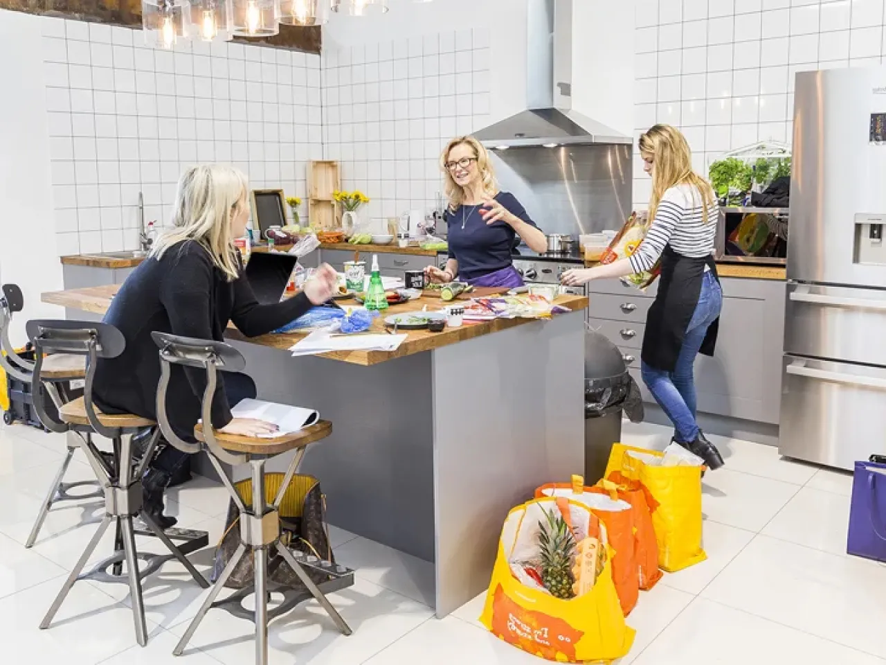 Three women in a modern kitchen preparing food, with grocery bags on the floor and various ingredients on the counter.