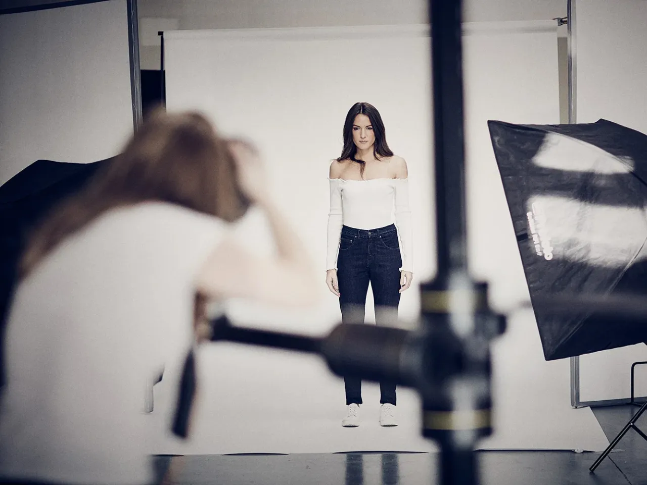 A woman in a white off-shoulder top and dark jeans posing for a photoshoot in a studio with lighting equipment.
