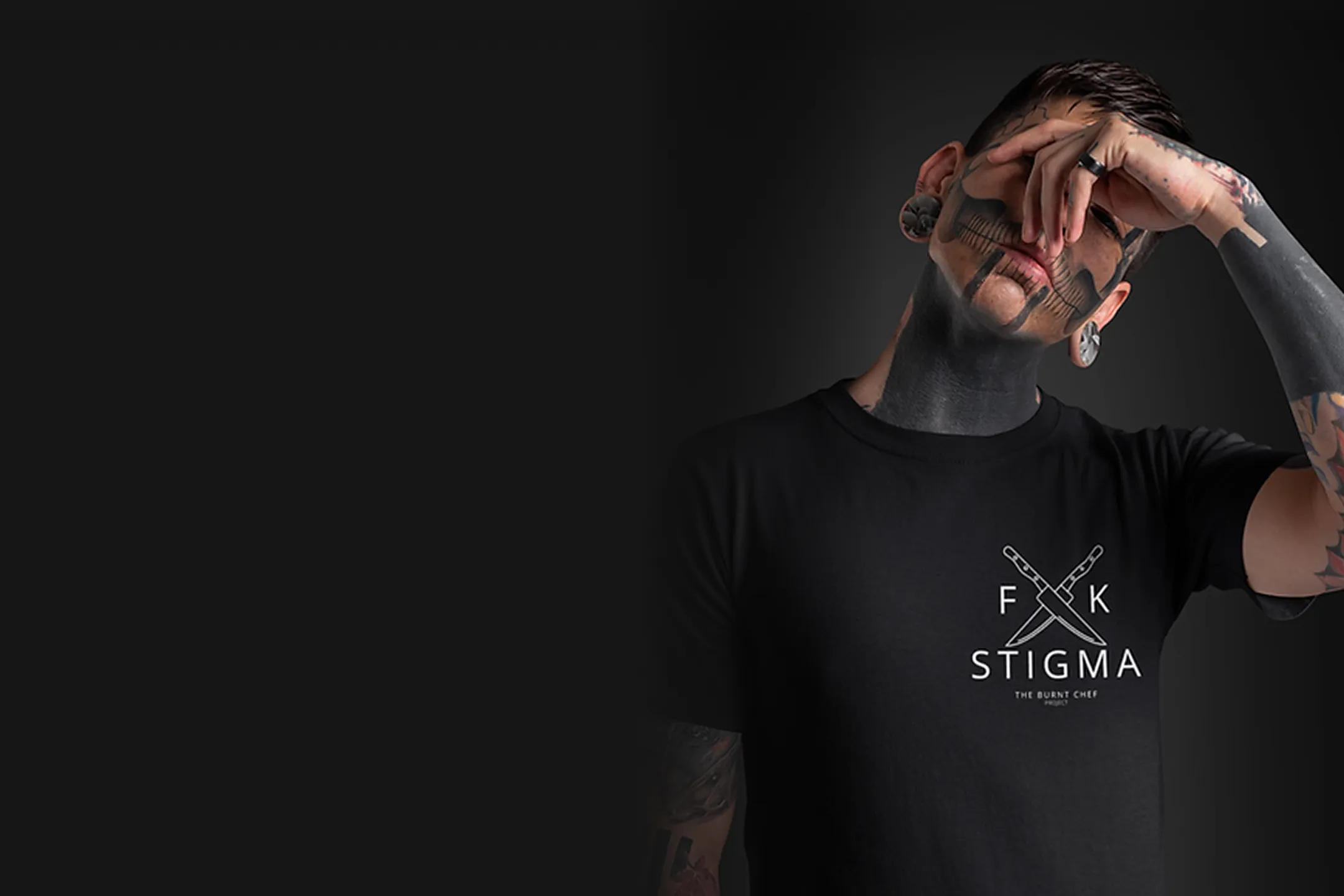 Tattooed man with skull face paint wearing black t-shirt that says 'F K STIGMA' with crossed knives logo.