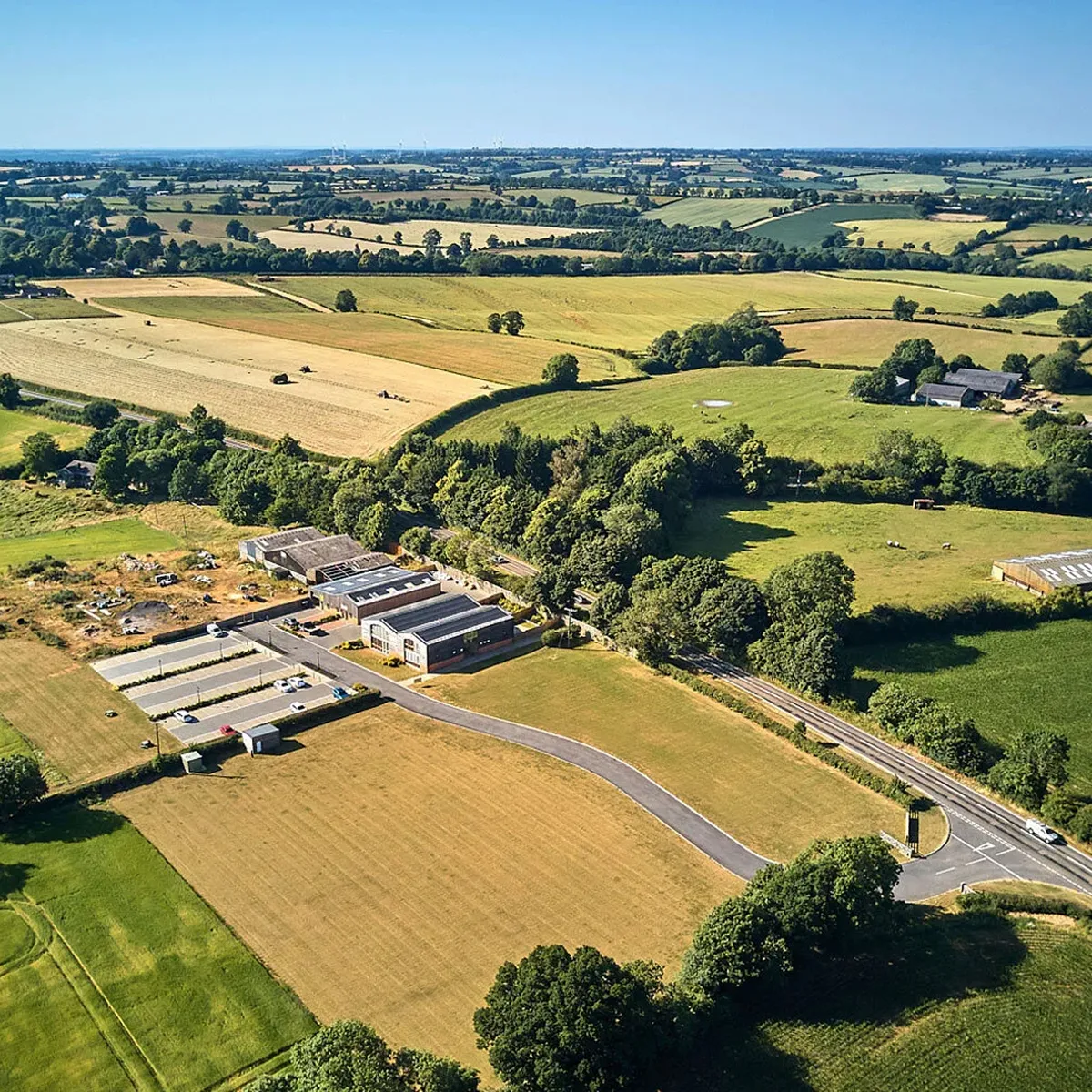 Aerial view of rural landscape with green and golden fields, scattered trees, farm buildings, and a curved road leading to a small parking area.
