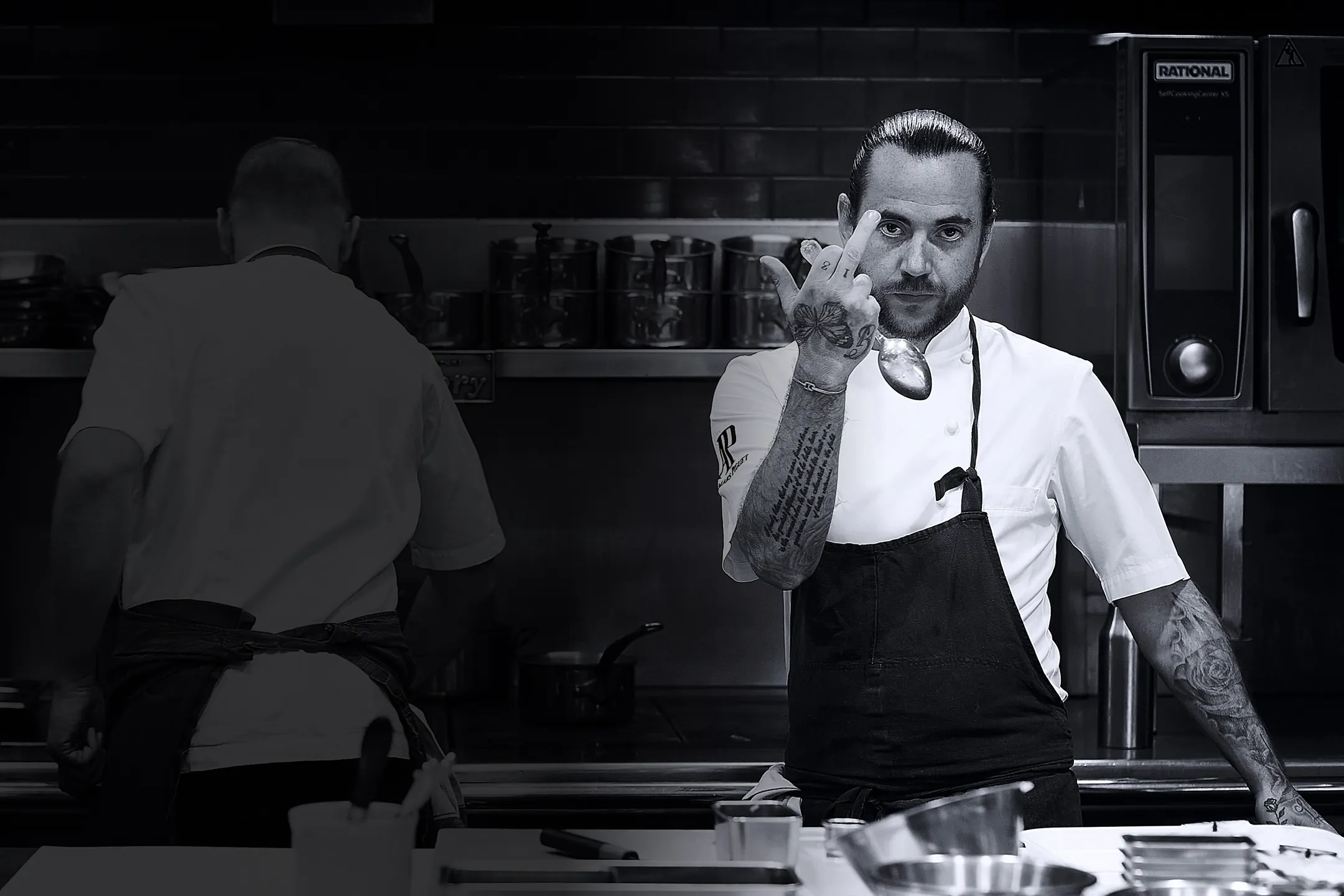 Tattooed male chef in white shirt and black apron holding a spoon and showing a middle finger in a professional kitchen.