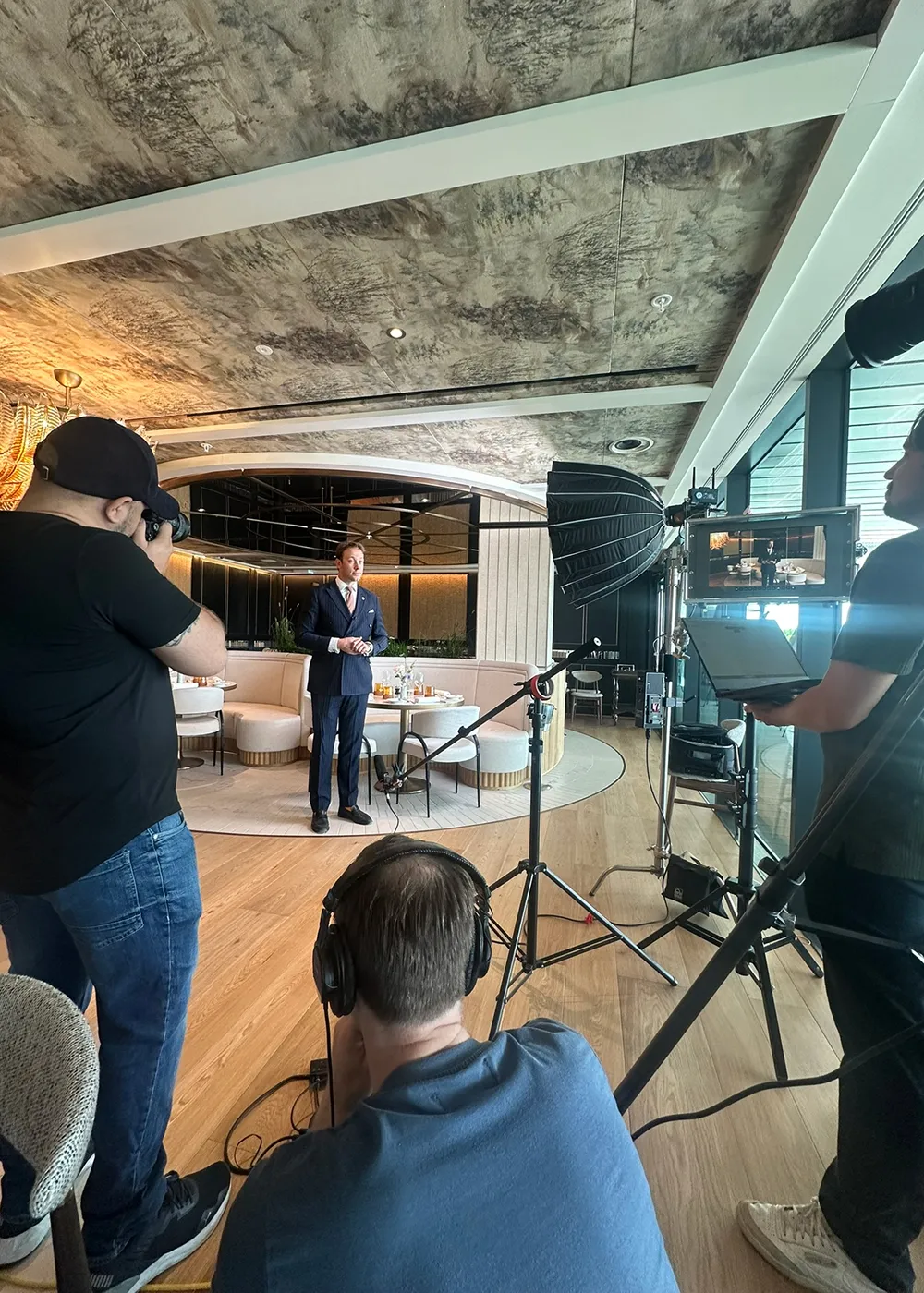 Man in a blue suit standing in a modern restaurant setting being photographed and recorded by a film crew with lighting and audio equipment.