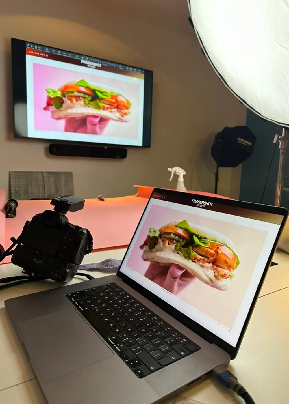 Photography studio setup with a laptop and TV screen displaying a close-up photo of a sandwich with lettuce, tomato, and grilled chicken.
