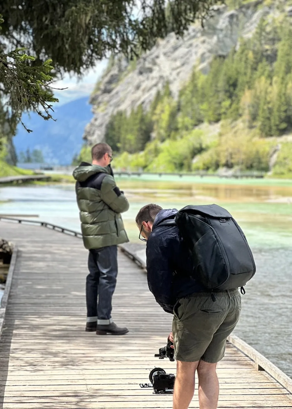 Two men on a wooden boardwalk by a river with trees and rocky hills in the background; one man is standing with arms crossed, the other is adjusting a camera on the boardwalk.