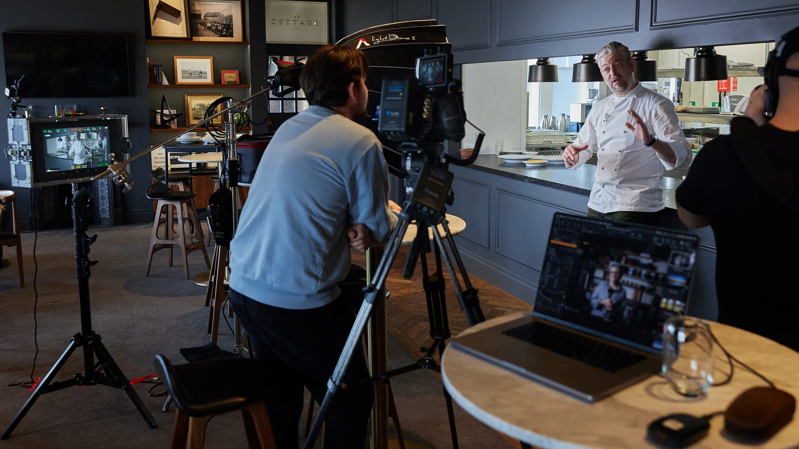 Video crew filming a chef in a white jacket speaking in a restaurant kitchen with camera and laptop in the foreground.