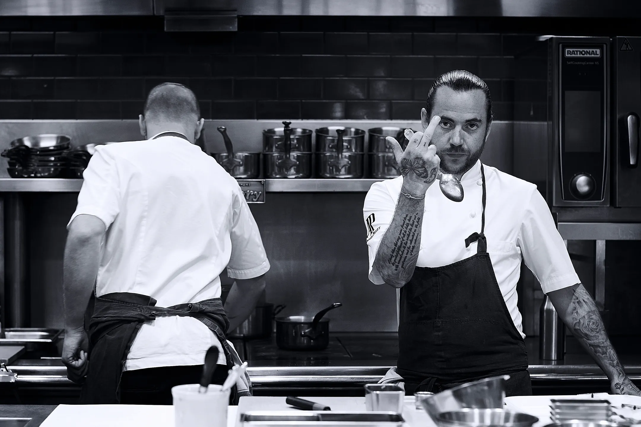 Two chefs working in a commercial kitchen, one facing away and the other showing a tattooed hand while holding a spoon and making a rude gesture.