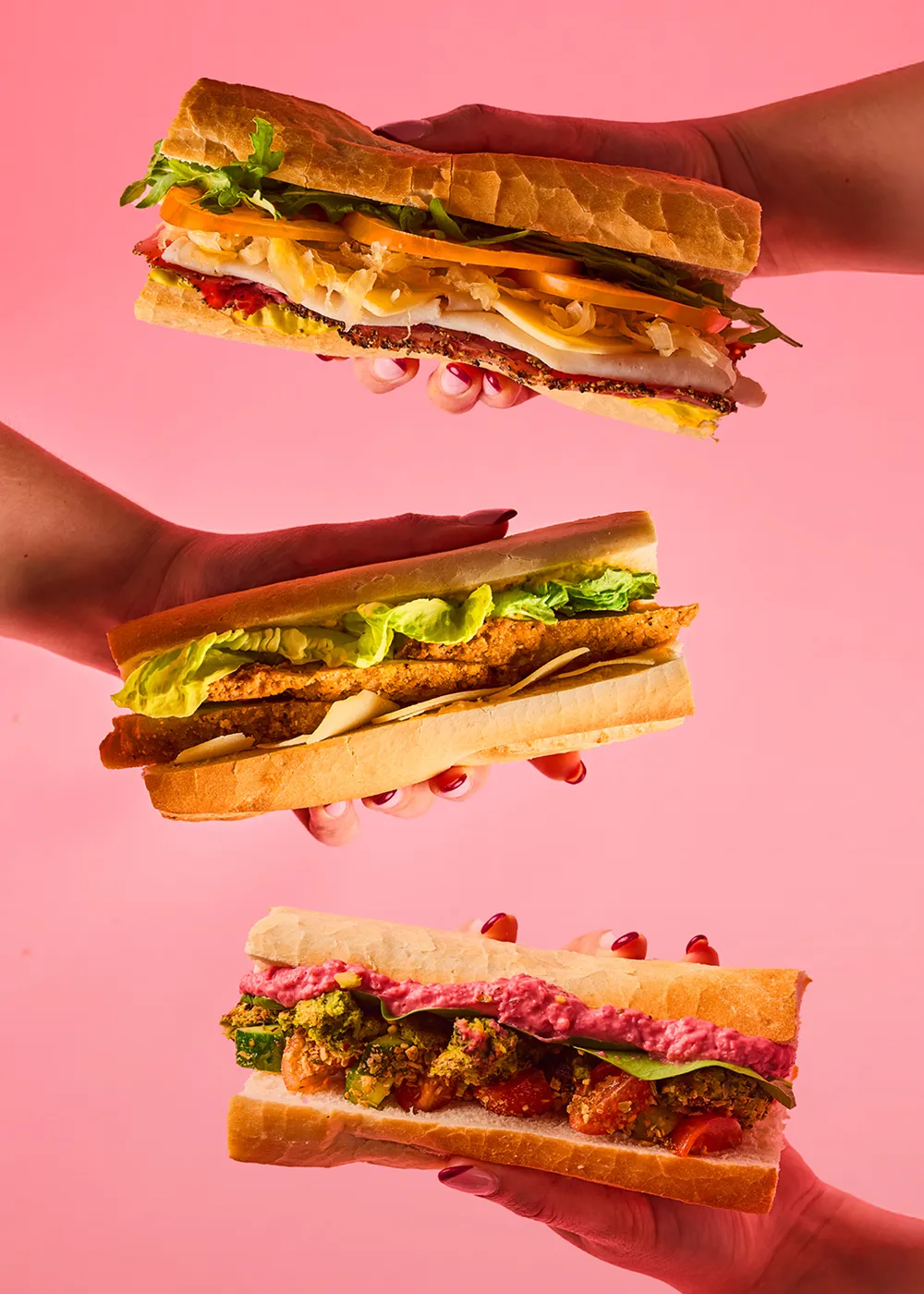 Three different sandwiches held by hands against a pink background, featuring deli meat with lettuce and tomato, breaded chicken with lettuce, and a veggie sandwich with pink hummus and greens.