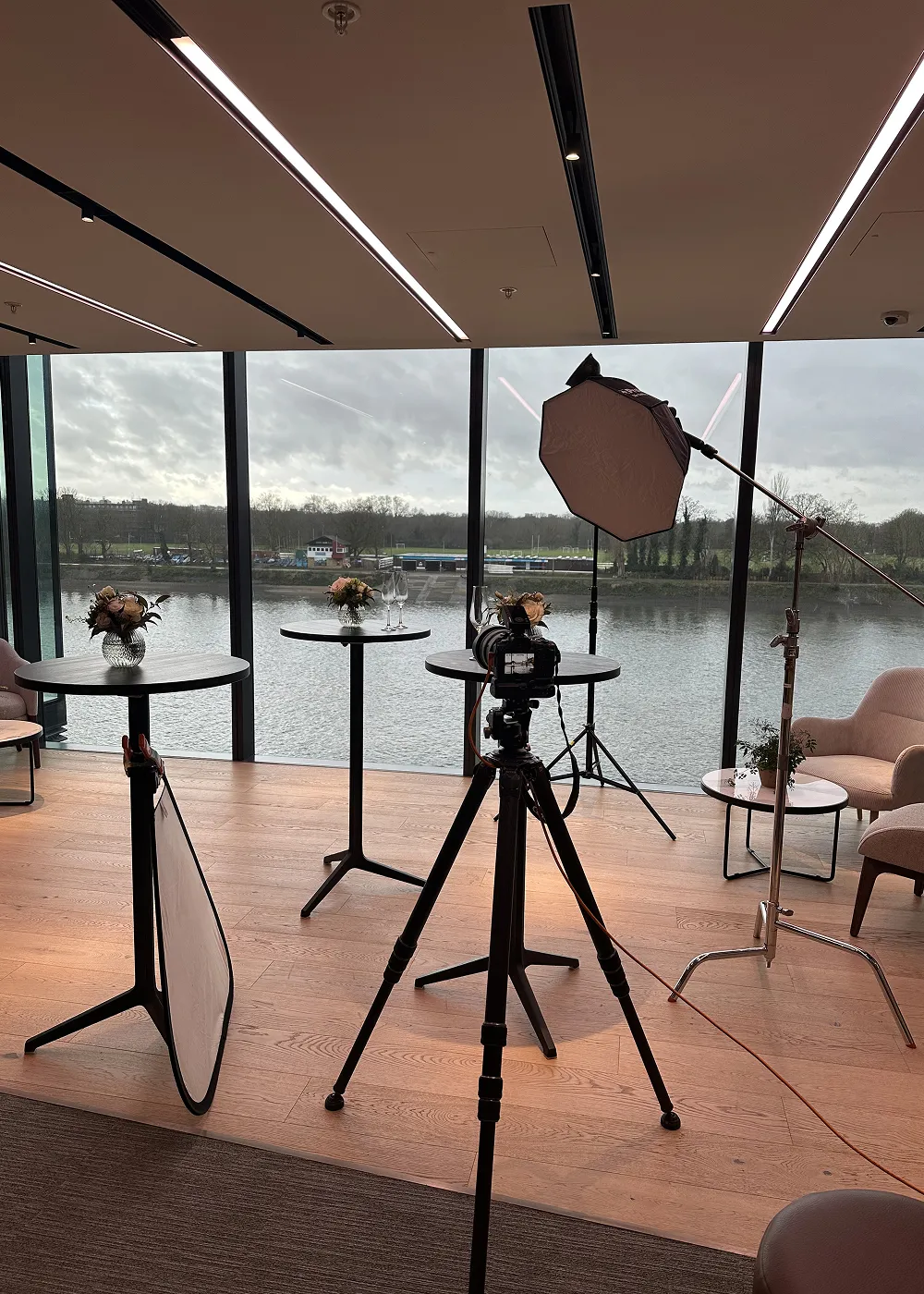 Indoor photography setup with camera on tripod, softbox light, reflector, and small tables with floral arrangements by large windows overlooking a river.