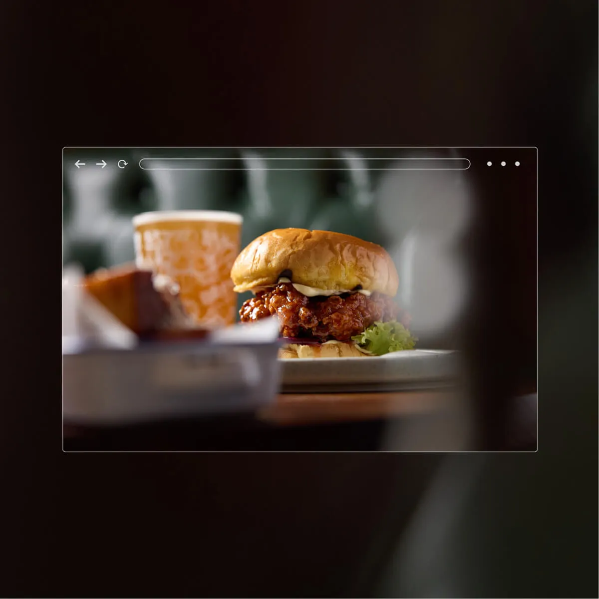 Close-up of a fried chicken sandwich with lettuce and sauce on a white plate, with a blurred drink and fries in the background.