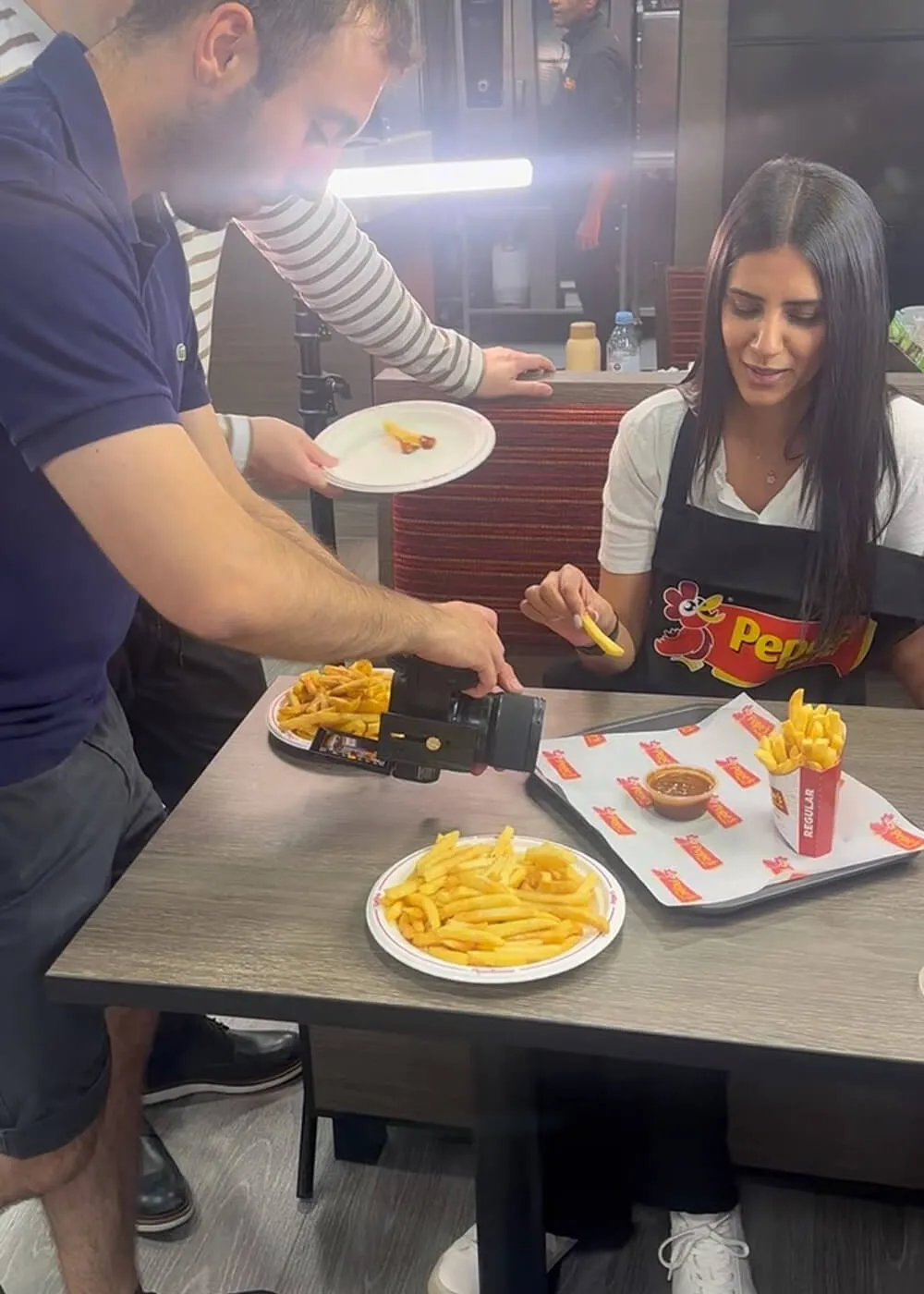 A woman wearing a Pepe's apron sitting at a table with plates and containers of French fries while a man takes close-up photos of the fries using a camera.