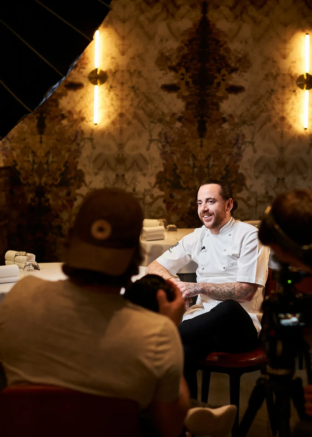 Chef with tattoos wearing white uniform smiling during a photoshoot in a warmly lit restaurant setting.