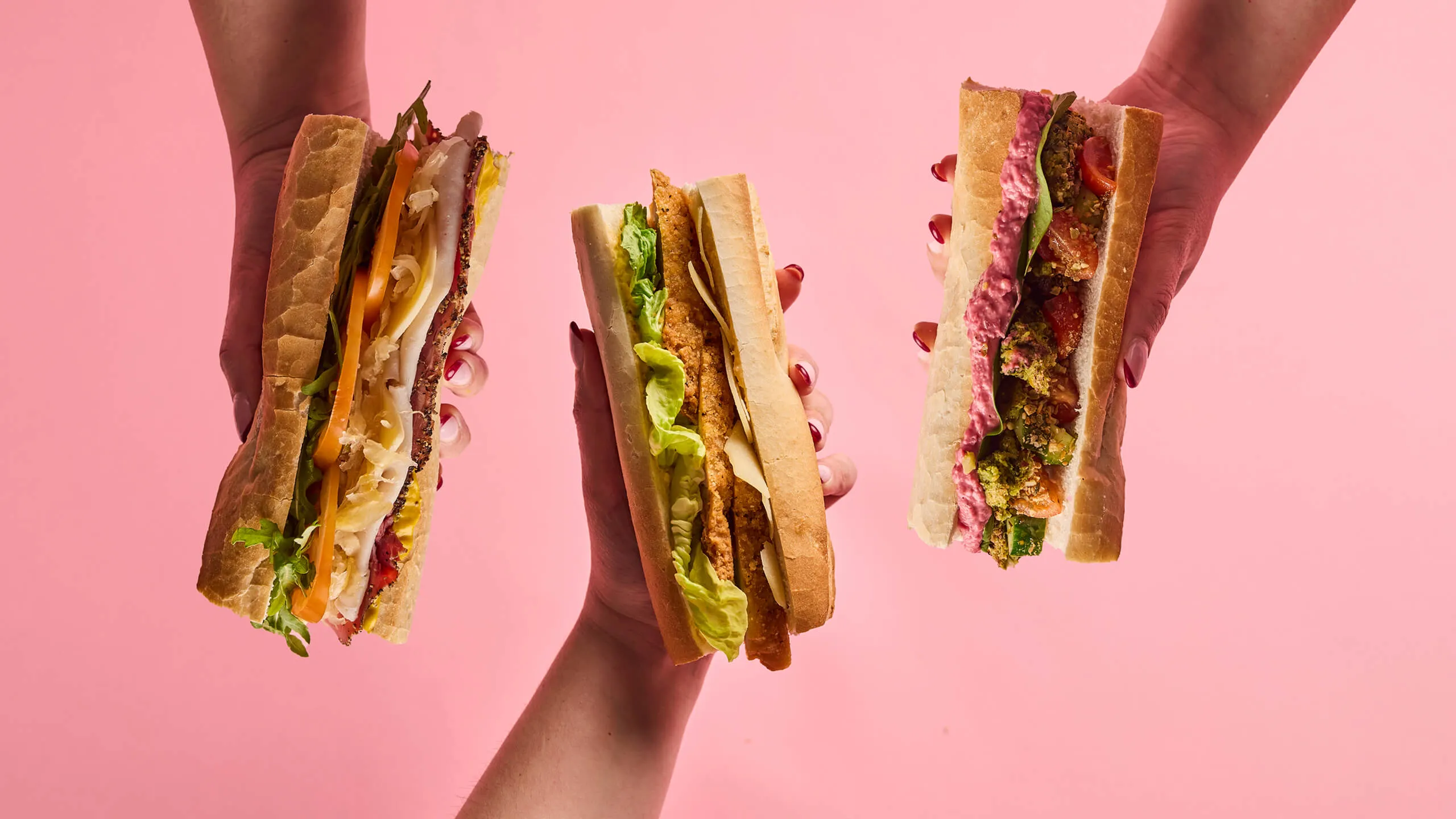 Three hands holding different sandwiches with various fillings against a pink background.