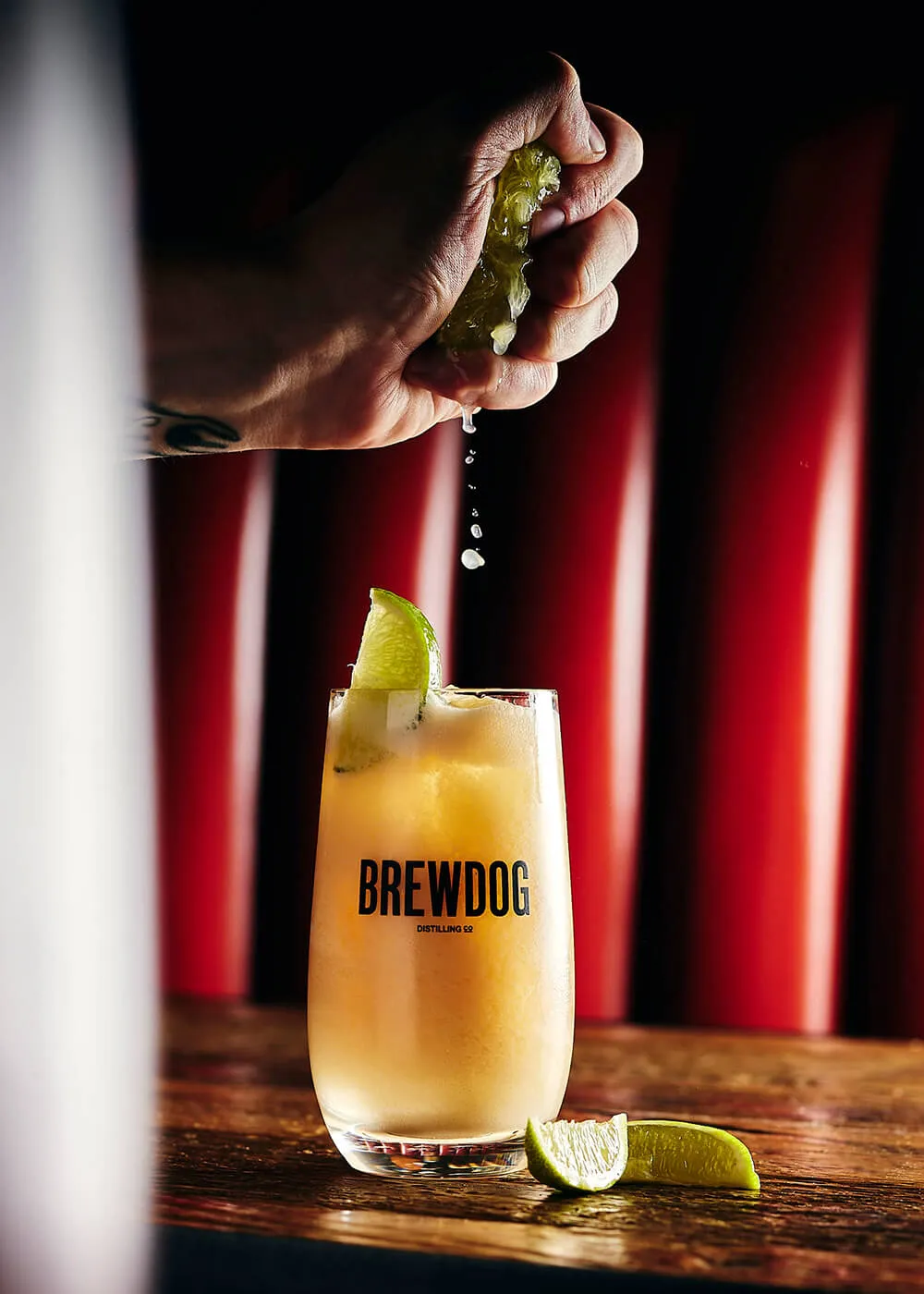 Hand squeezing lime juice into a BrewDog cocktail glass filled with ice and garnished with a lime wedge, on a wooden surface.