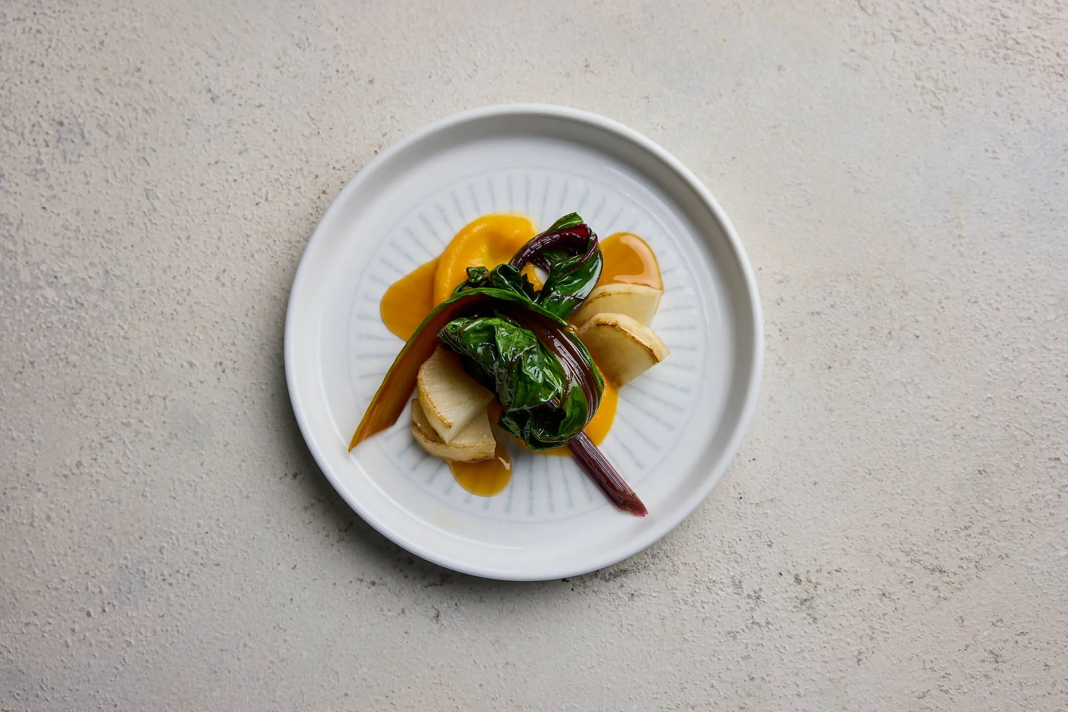 Plate with cooked green leafy vegetable, sliced root vegetables, and orange sauce on a light surface.