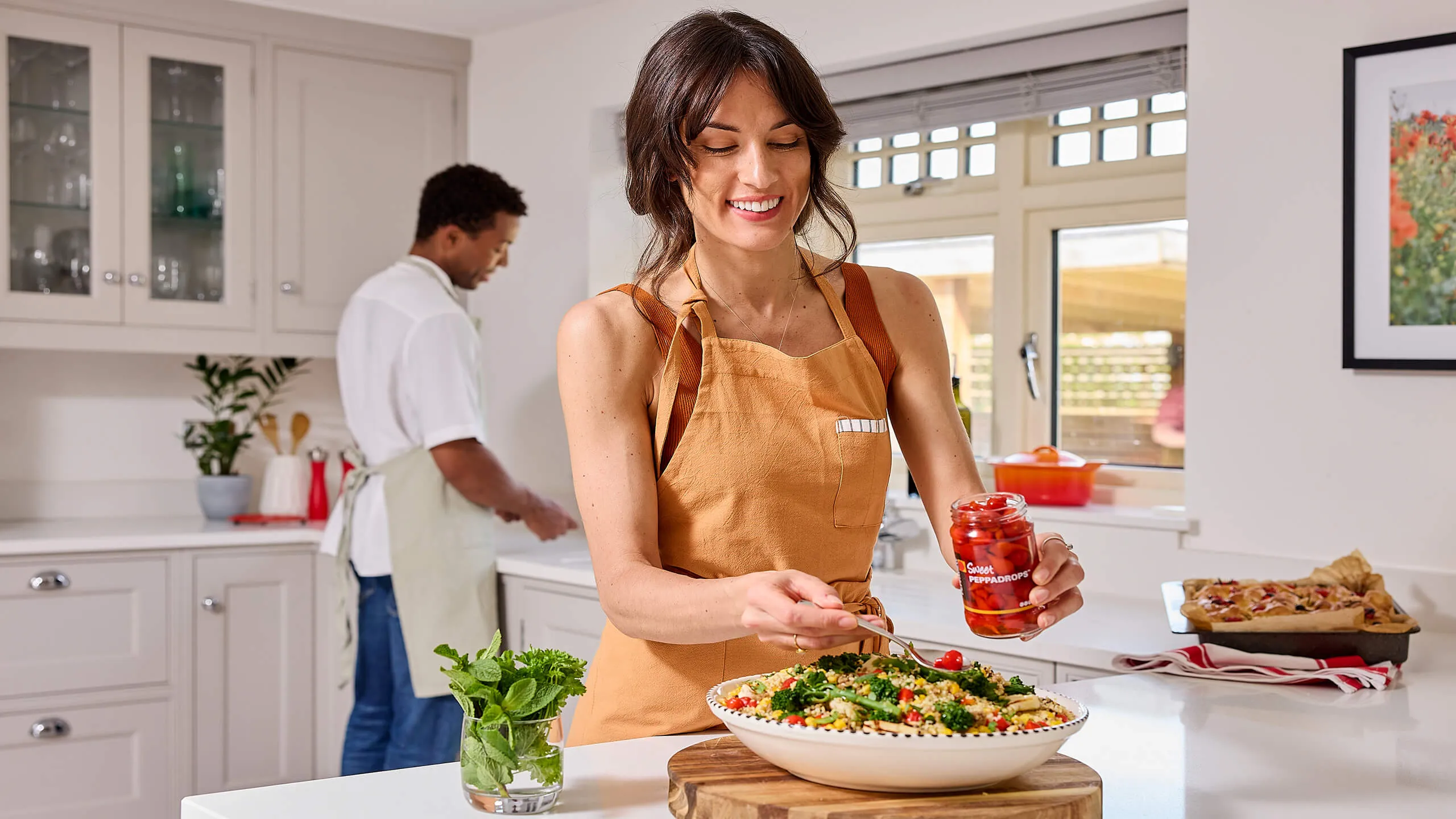Woman in a brown apron adding red Peppadrops to a large bowl of couscous salad in a bright kitchen, with a man cooking in the background.
