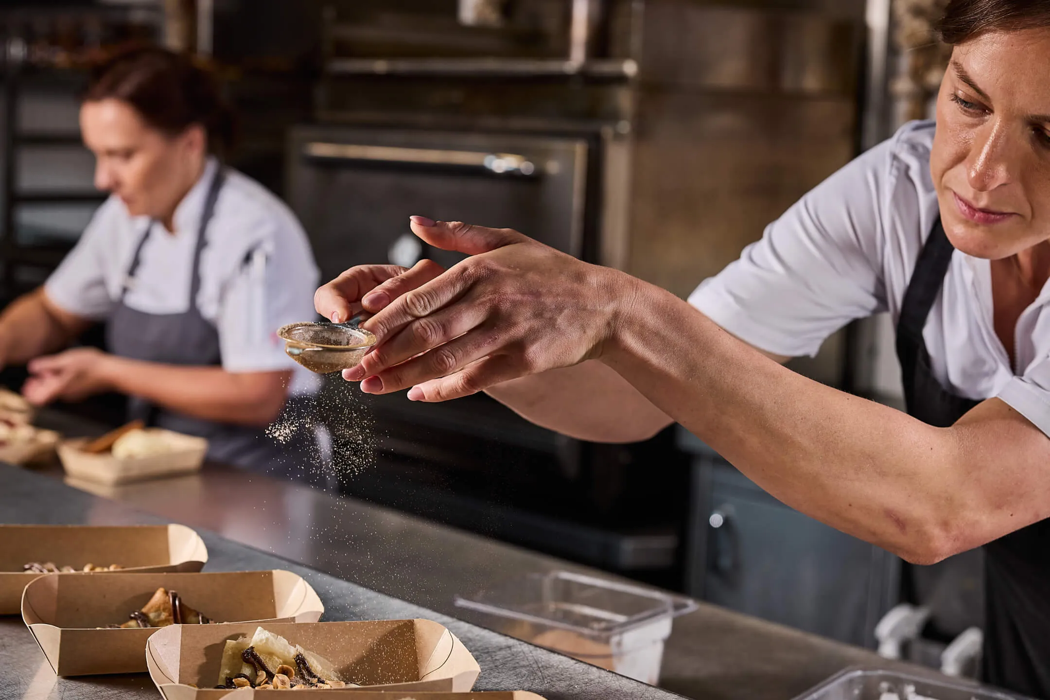 Chef dusting powdered ingredient over trays of prepared food in a professional kitchen.