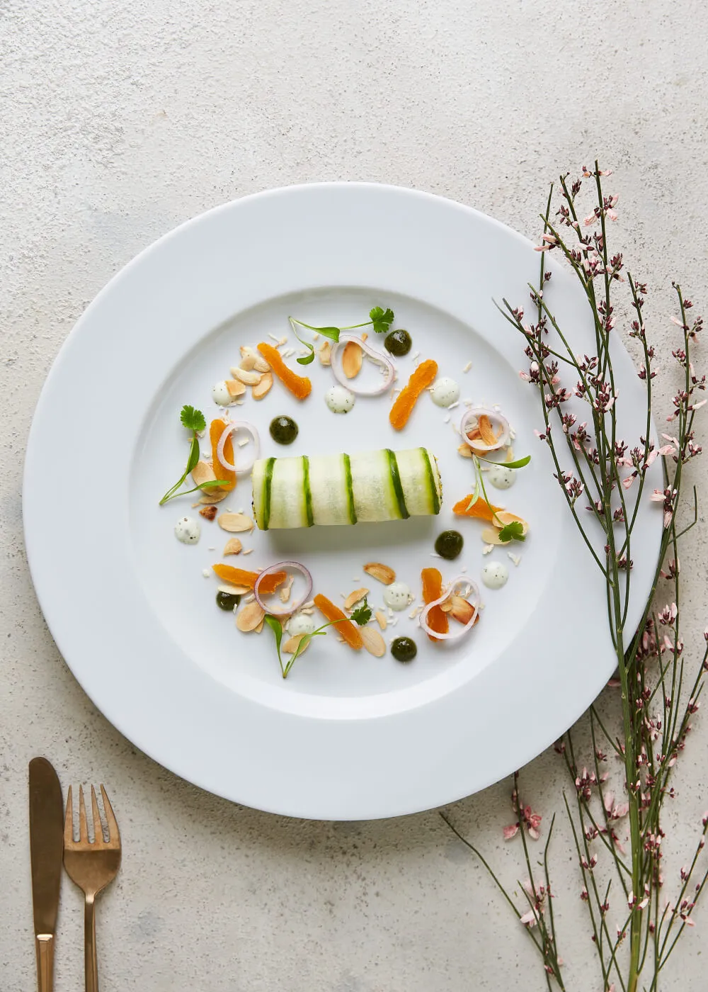 White plate with a cylindrical cucumber-wrapped dish garnished with mandarin orange segments, almond slices, thin onion rings, green herbs, and small dollops of green and white sauce, accompanied by a fork and knife, and pink flowering branches on the side.