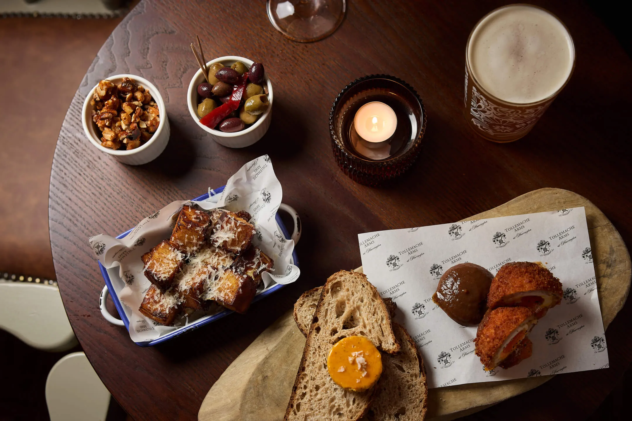 Wooden table with a lit candle, a pint of beer, small bowls of nuts and olives, fried bread cubes with grated cheese, slices of bread, and a halved Scotch egg with brown sauce on branded paper.