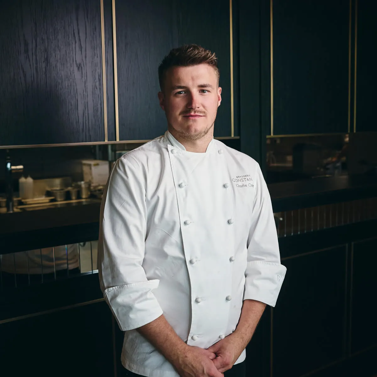 Chef wearing a white double-breasted jacket standing in a modern kitchen with dark cabinets.