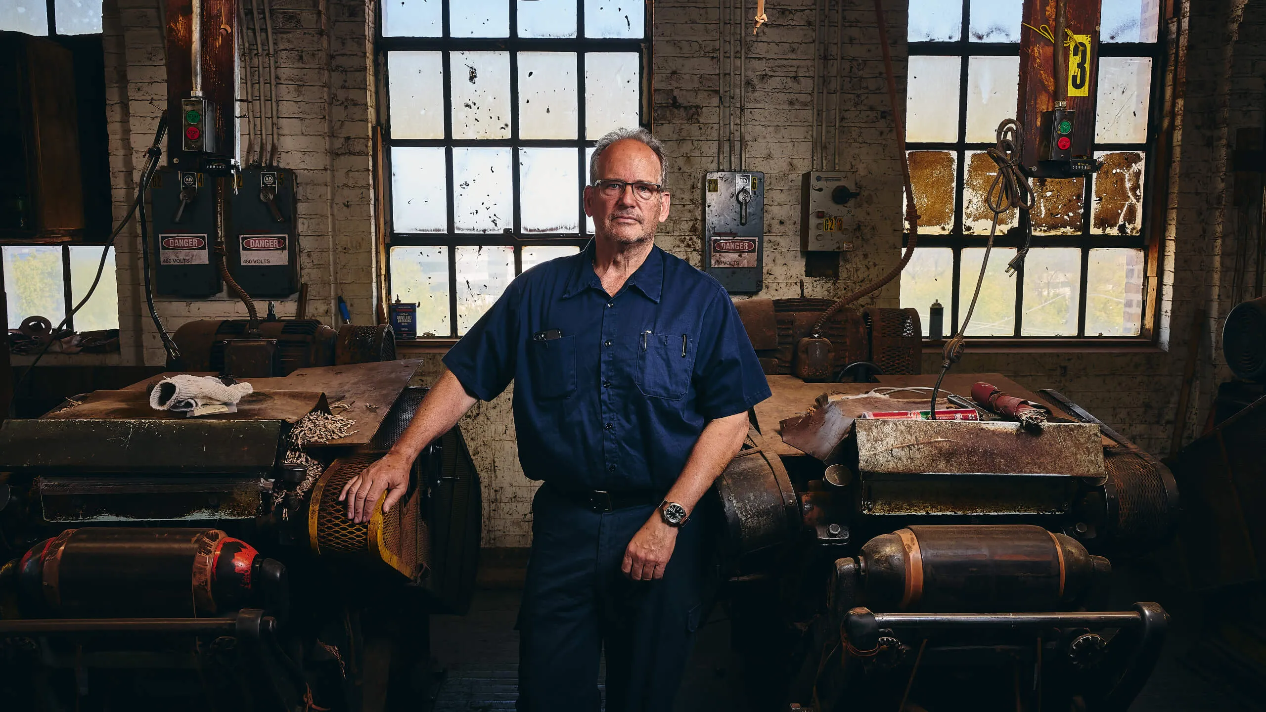 Man in navy workwear standing in an old industrial workshop with large machinery and dirty windows behind him.