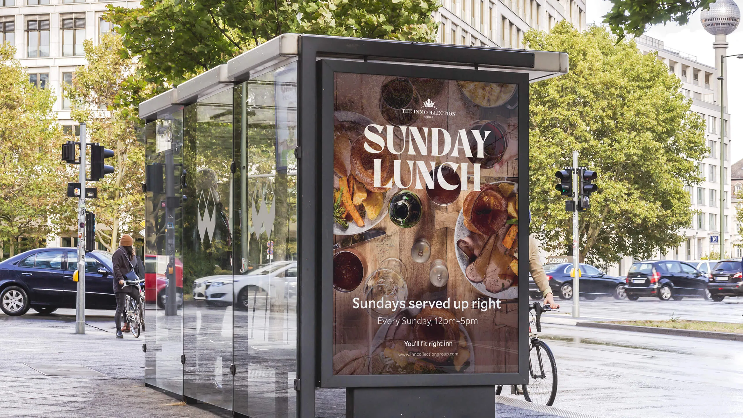 Bus stop digital display showing an advertisement for The Inn Collection's Sunday Lunch with plates of roasted meat, vegetables, and wine glasses on a wooden table.