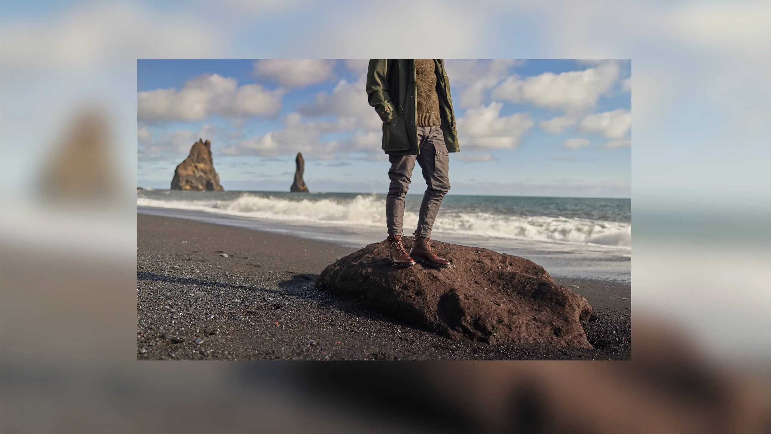 Person wearing brown leather boots standing on a large rock on a black sand beach with ocean waves and rock formations in the background.