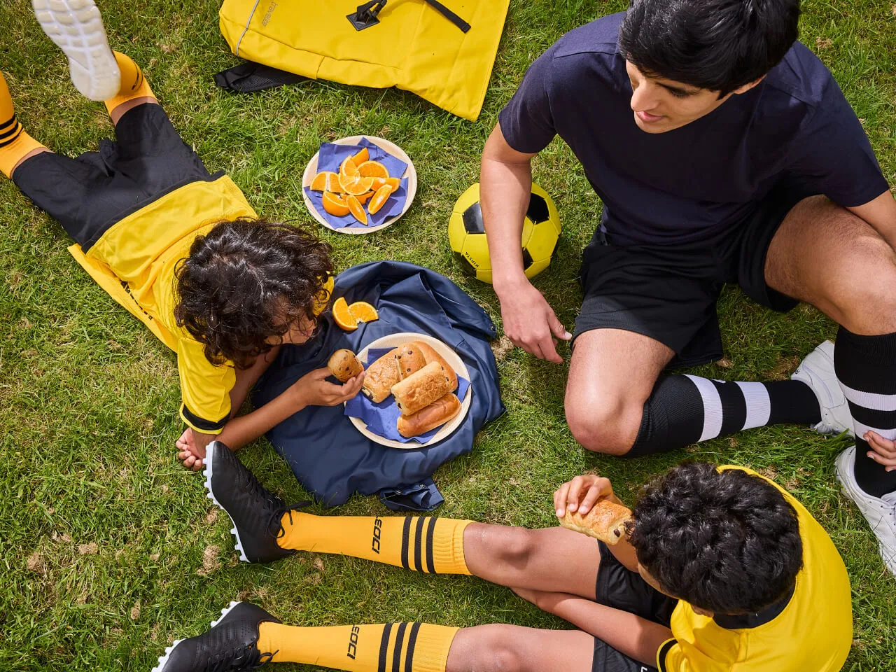 Two boys in yellow football kit and a coach sitting and lying on grass having snacks and orange slices beside a yellow and black soccer ball.