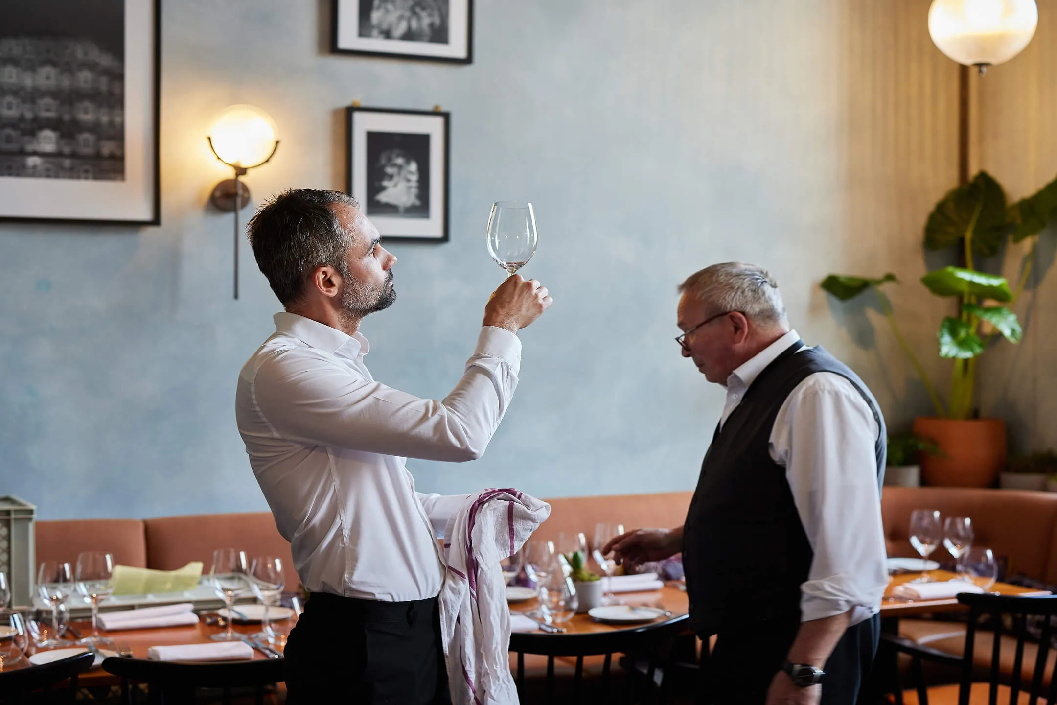A waiter inspects a wine glass while another stands nearby in a restaurant dining area.